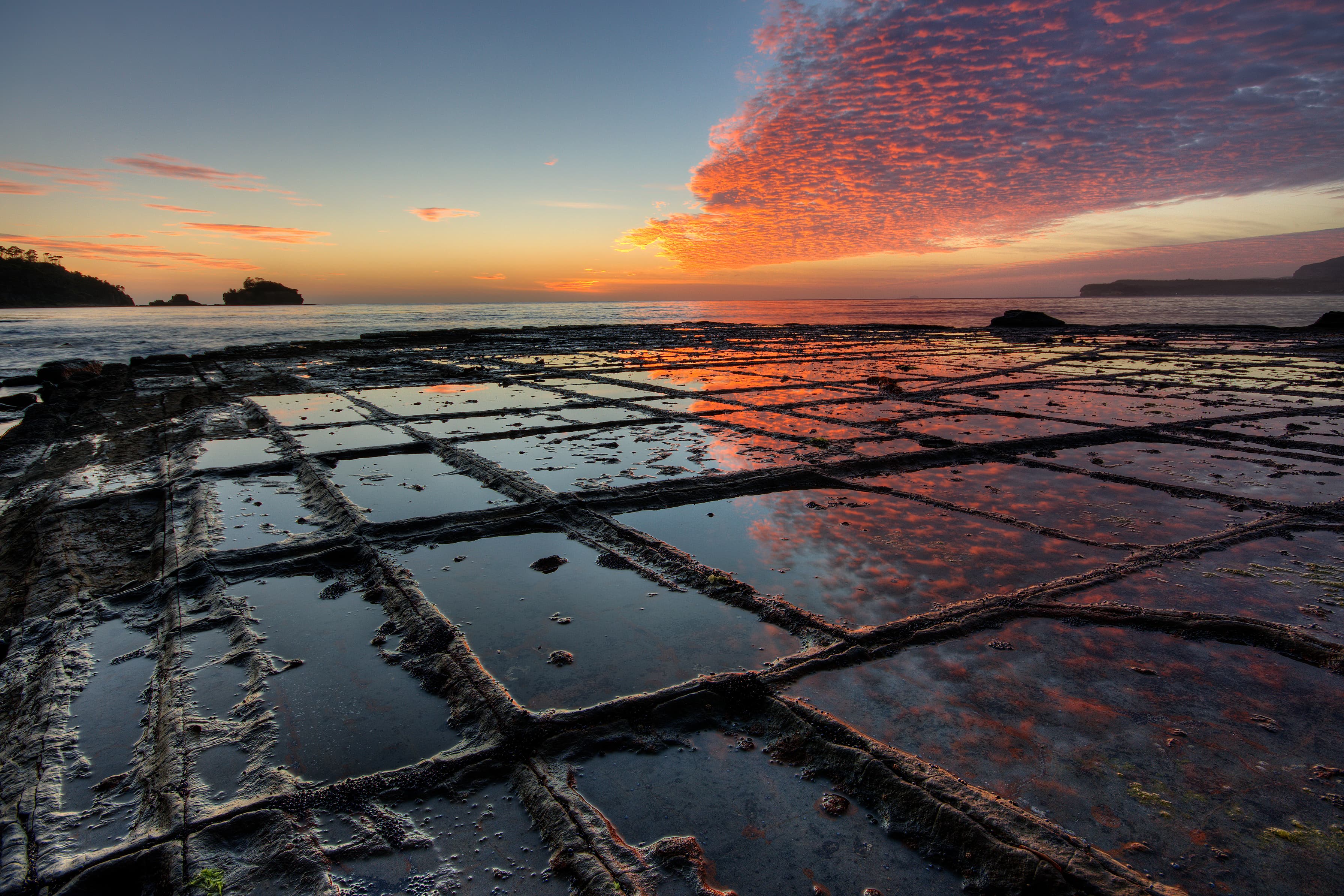 Sunrise, “Tessellated Pavement”, Eaglehawk Neck, Tasman Peninsula, Tasmania, Australia. The „Tesselated Pavement“ is the result of an orthogonal joint pattern in the rock. On the picture it shows the so called “pan formation”, where the rock in the immediate vicinity of the joints is more resistant to erosion than the rock that is more distant to the joints, This is due to alterations of the rock along the joints by hydrothermal (or similar) solutions when the rock was still buried deeply below the surface millions of years ago. When no alterations or alterations that lower the erosional resistivity have taken place in the geological past, the rock along the joints will erode faster than the rock that is more distant to the joints. In that case the so called “loaf formation” of “Tessellated Pavement” will form.